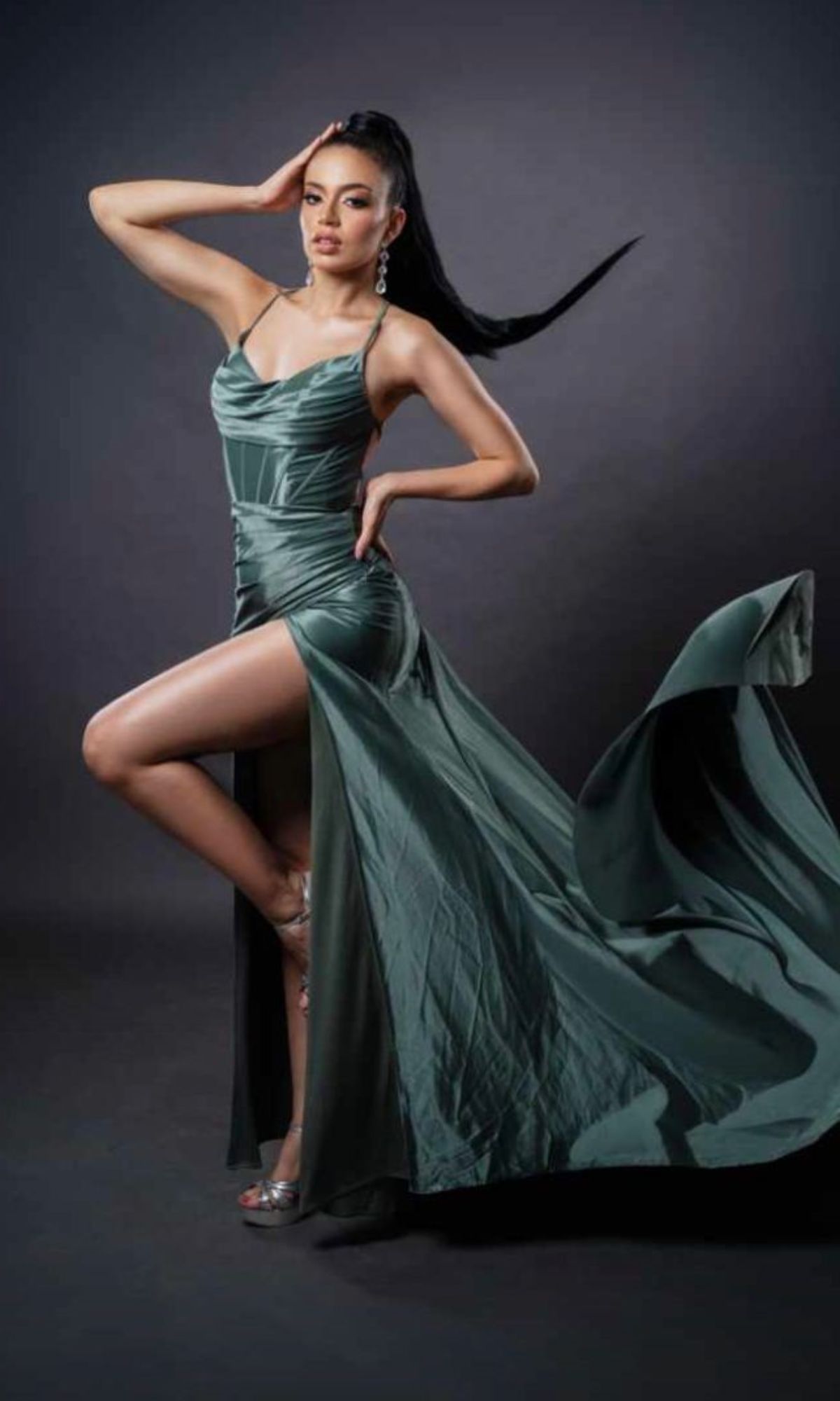 Woman posing in a studio against a dark background, wearing the Cowl-Neck Long Prom Dress: Chicas C326—a flowing green strappy-back gown with a high slit—paired with silver heels and statement earrings.